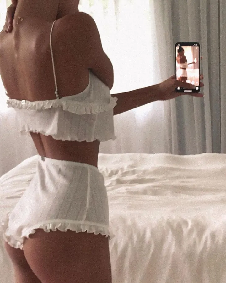 Femme en lingerie blanche frangée, prenant un selfie devant un lit blanc, tenant un smartphone.