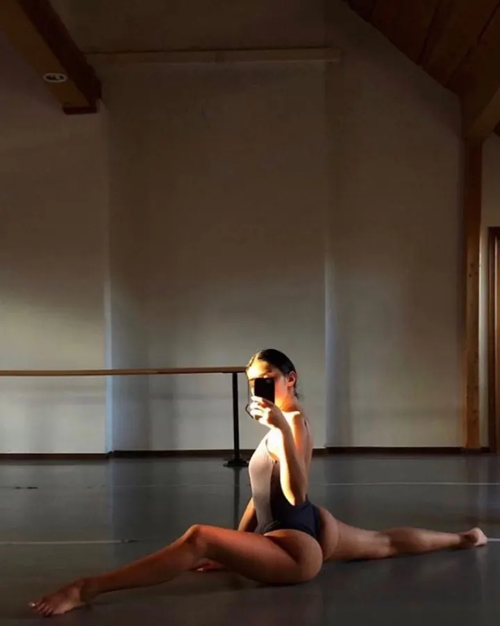 Femme en justaucorps noir, prenant un selfie dans une salle de danse, en position de split.
