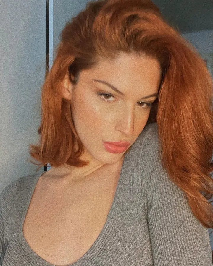 Femme aux cheveux roux, en pull gris, regardant vers le bas avec un air pensif.