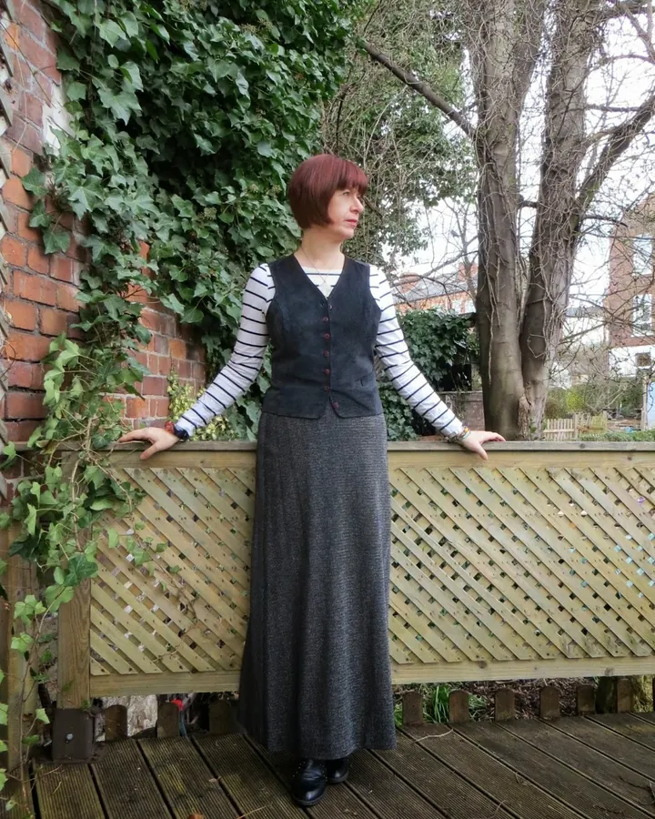 Femme en robe longue grise et gilet noir, debout sur une terrasse en bois, appuyée contre une clôture en treillis, avec des feuilles de lierre sur un mur de briques.