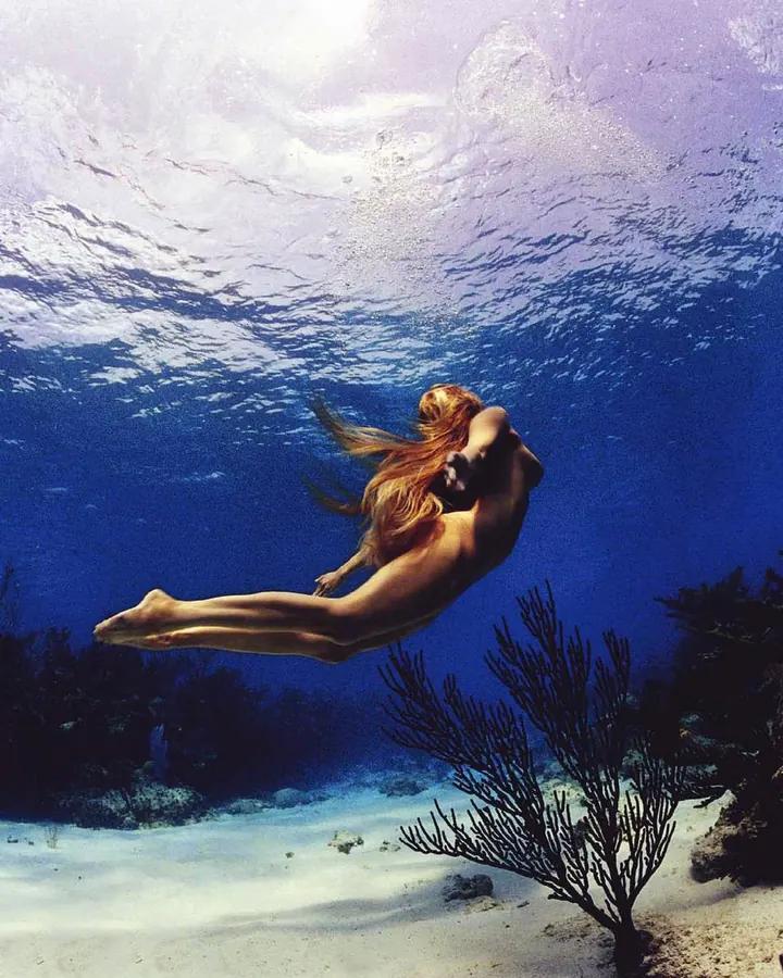 Femme aux cheveux blonds en maillot de bain, flottant sous l'eau, avec des coraux en arrière-plan.