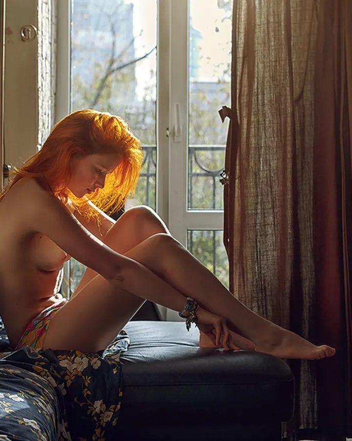 Femme nue, assise sur un lit, les cheveux roux éclairés par la lumière du soleil, les jambes croisées, regardant vers le bas, ambiance intime et chaleureuse.