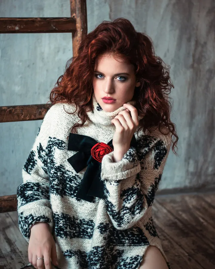 Femme européenne aux cheveux roux, portant un pull à motifs noir et blanc avec un ruban noir et une rose rouge, assise sur une chaise en bois.