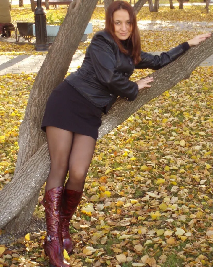 Femme en tenue noire, collant noir et talons hauts, tenant une petite cravache, posant en extérieur.