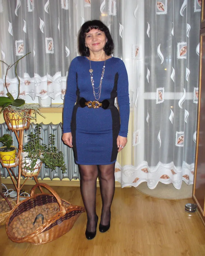 Femme aux cheveux noirs portant une robe bleue avec des bandes noires, un collier long, et des bas noirs, debout dans une pièce avec des plantes et un panier de noix.