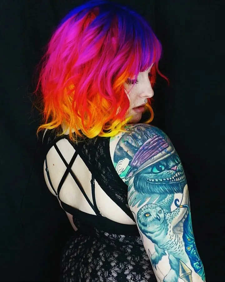 Femme aux cheveux multicolores (rose, rouge, orange, jaune) portant une robe noire en dentelle, tatouage visible sur le bras gauche, profil sur fond noir.