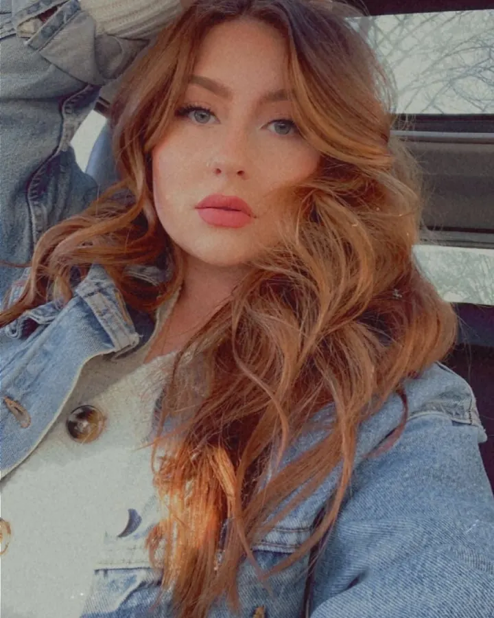 Femme aux cheveux blonds ondulés portant une veste en jean, regardant l'appareil photo avec un air pensif, dans une voiture.