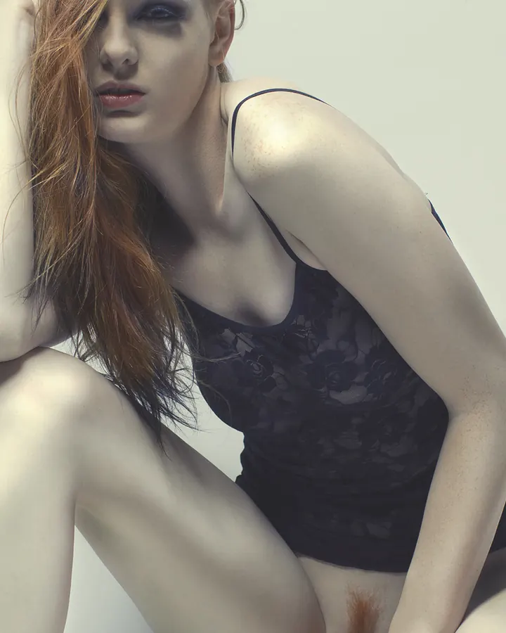 Femme européenne aux cheveux roux, en lingerie noire, tenant son cou, cheveux flottant, lumière douce, regardant la caméra.