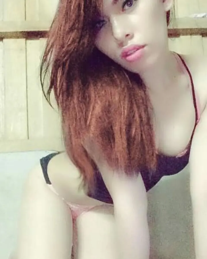 Femme latino aux cheveux roux, en t-shirt blanc, prenant un selfie dans un miroir, main sur le ventre, sous-vêtement noir visible.