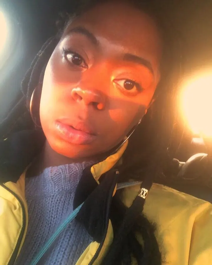 Femme africaine aux cheveux longs noirs, en t-shirt noir, regardant la caméra de face, visage net, cheveux tressés avec mèches blondes, lumière chaude.