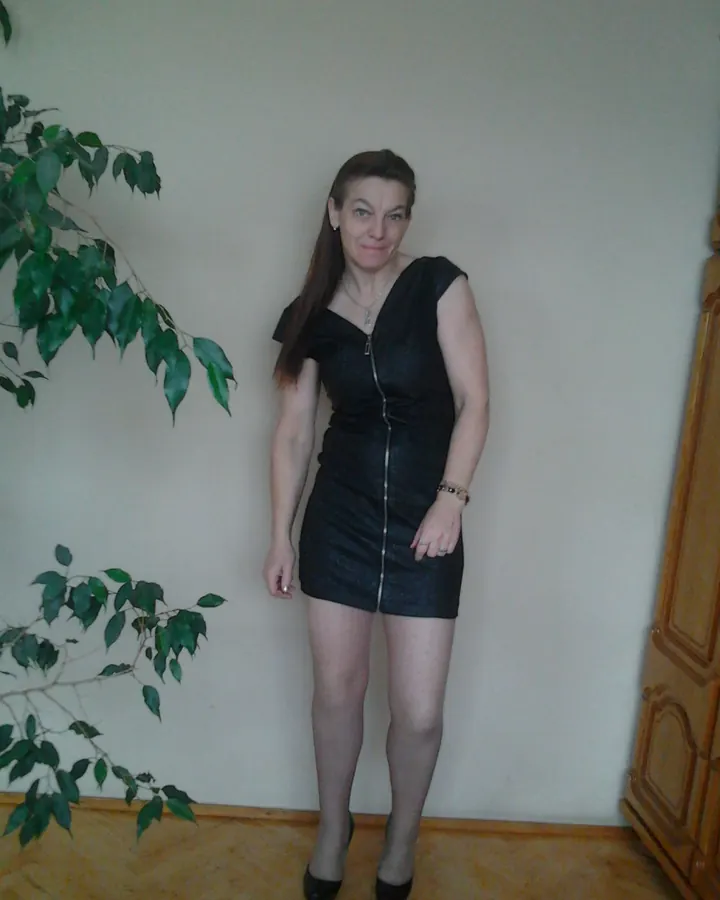 Femme aux cheveux bruns portant une robe noire à zip, debout près d'une plante verte et d'un meuble en bois.
