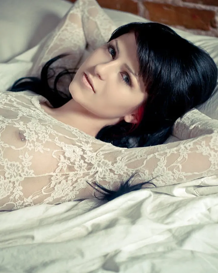Femme aux cheveux bruns-noirs en lingerie blanche, posant de profil contre un mur bleu.