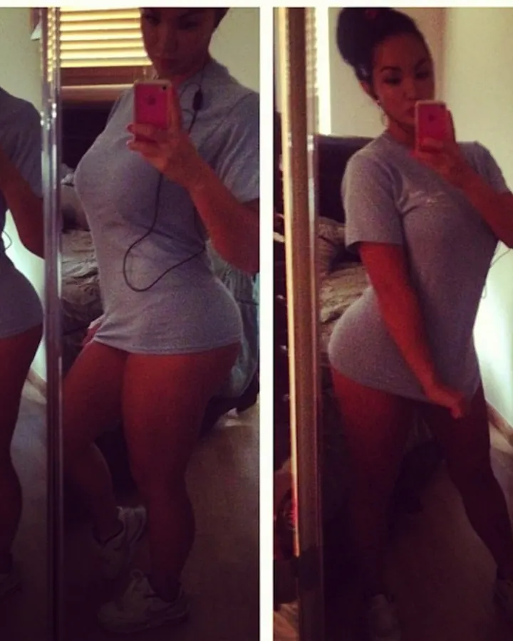 Femme aux cheveux bruns-noirs en t-shirt gris, prenant un selfie devant un miroir dans une chambre.