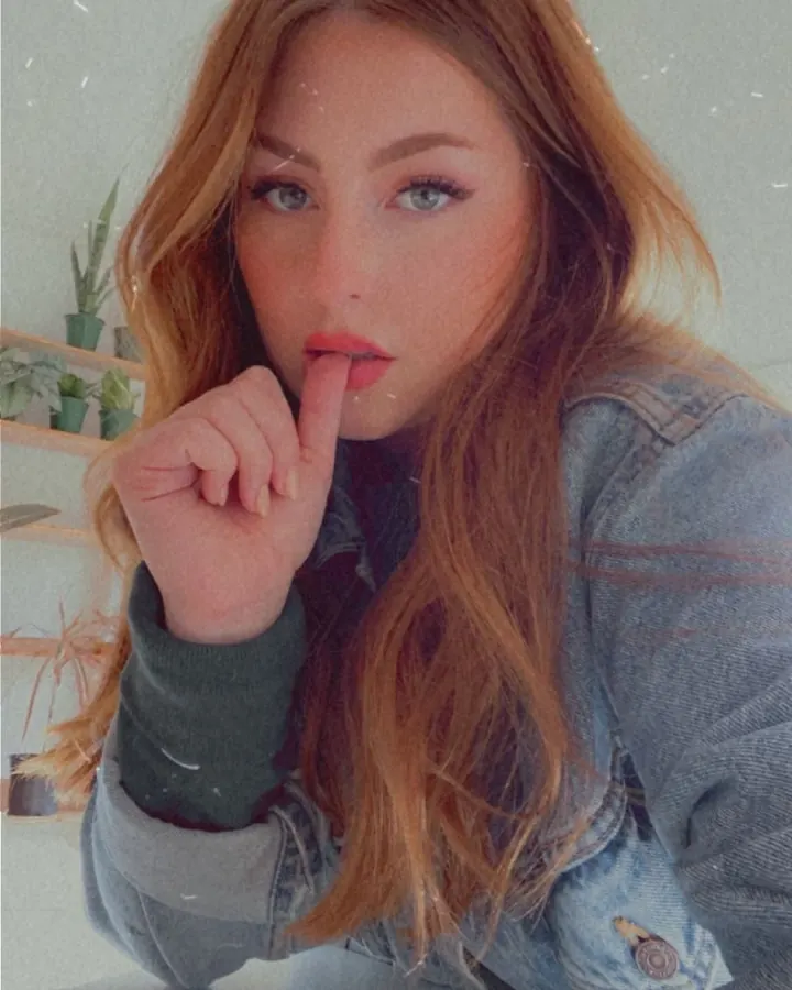 Femme aux cheveux roux portant un jean et un pull vert, posant avec un doigt sur les lèvres.