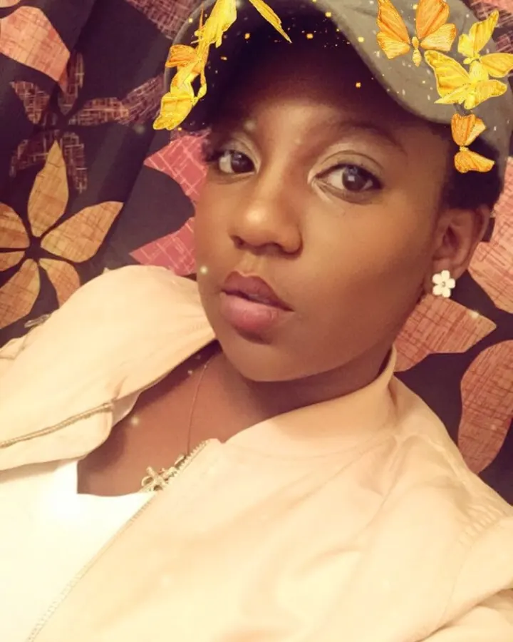 Femme africaine aux cheveux courts, portant une casquette grise ornée de papillons dorés, vêtue d'une veste beige, regard fixe et sérieux.