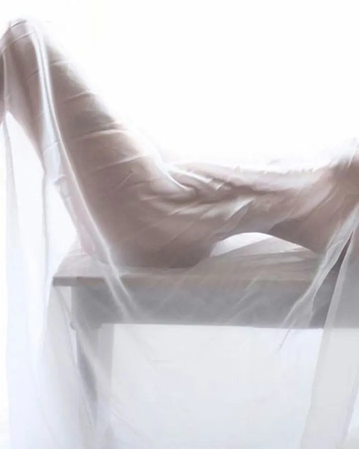 Femme nue en transparence blanche, allongée sur une table, les jambes levées, main dans les cheveux