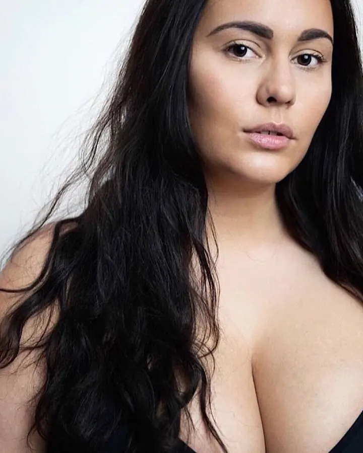 Femme aux cheveux noirs en haut noir décolleté, regardant l'appareil photo avec un regard intense.