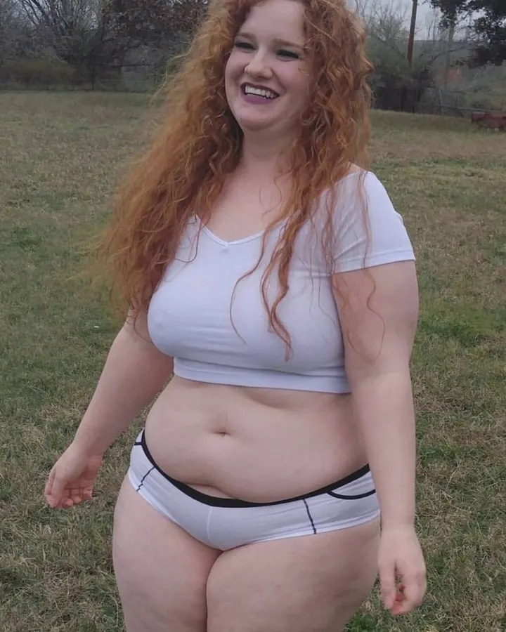 Femme aux cheveux roux bouclés, en haut blanc et culotte blanche, souriant largement dans un champ herbeux.