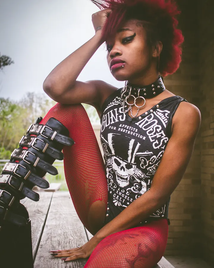 Femme aux cheveux roux, en tenue punk avec choker, collants rouges et bottes noires, posant sur un banc en bois.