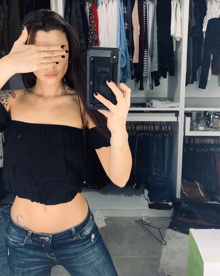 Femme en lingerie blanche ajourée, debout dans une chambre avec rideau et bureau.