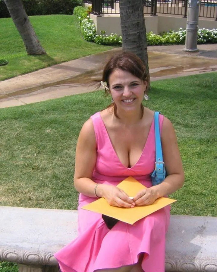 Femme souriante en robe rose vif, assise sur un banc en extérieur avec des palmiers en arrière-plan.